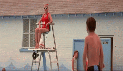 A Woman In A Red Bathing Suit Is Sitting On A Lifeguard Tower Next To A Boy . GIF