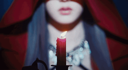 A Woman In A Red Cape Is Holding A Lit Candle In Front Of Her Face GIF