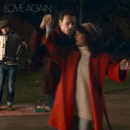 A Woman In A Red Coat Is Dancing With A Man Playing An Accordion And The Words Love Again Are Above Them GIF