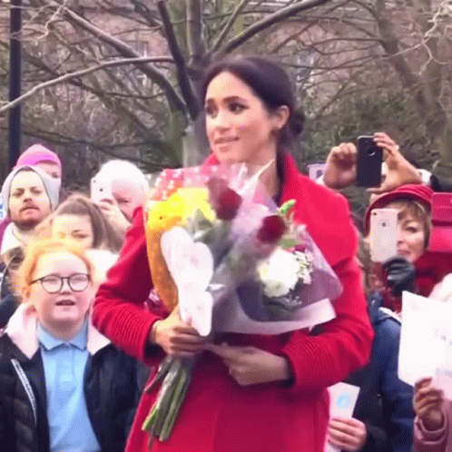 A Woman In A Red Coat Is Holding Flowers In Front Of A Crowd GIF