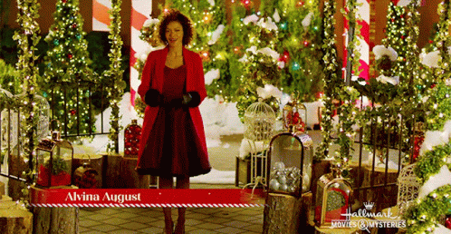 A Woman In A Red Coat Is Standing In Front Of Christmas Decorations And A Sign That Says " Alvina August " GIF
