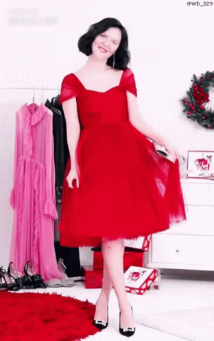 A Woman In A Red Dress And Black Heels Is Standing In Front Of A Wreath . GIF