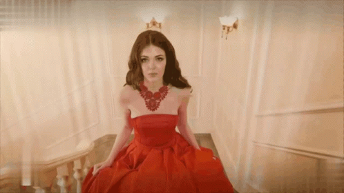 A Woman In A Red Dress And Necklace Is Sitting On A Set Of Stairs GIF