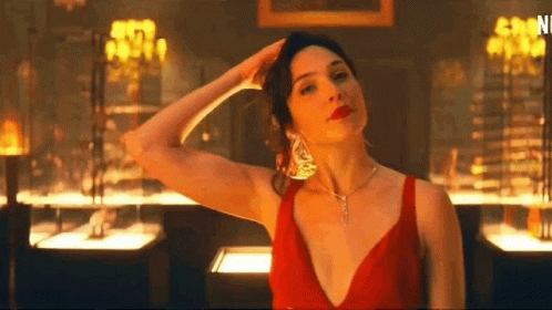 A Woman In A Red Dress And Necklace Is Standing In A Room With Her Hand On Her Head . GIF
