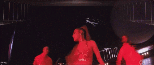 A Woman In A Red Dress Is Dancing On A Stage With A Man In A Red Shirt Behind Her GIF