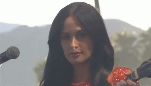 A Woman In A Red Dress Is Singing Into A Microphone With Mountains In The Background GIF