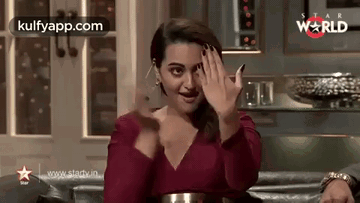 A Woman In A Red Dress Is Sitting On A Couch And Waving Her Hands . GIF