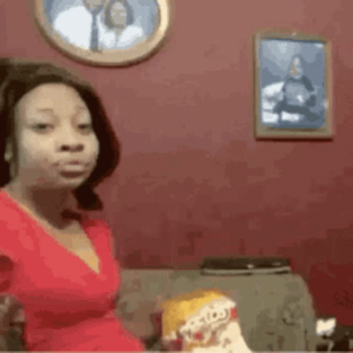 A Woman In A Red Dress Is Sitting On A Couch With A Picture Of A Man On The Wall Behind Her . GIF