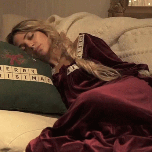 A Woman In A Red Dress Is Sleeping On A Bed Next To A Green Merry Christmas Pillow GIF