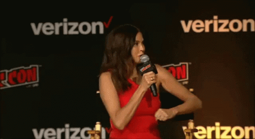 A Woman In A Red Dress Is Speaking Into A Microphone In Front Of A Verizon Sign GIF