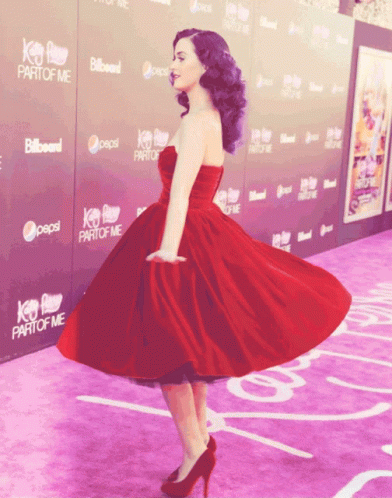 A Woman In A Red Dress Is Standing On A Purple Carpet With Pepsi And Billboard Signs Behind Her GIF
