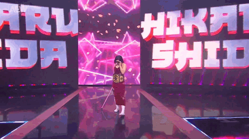 A Woman In A Red Dress Is Standing On A Stage In Front Of A Large Screen That Says ' Nikki Shida ' GIF