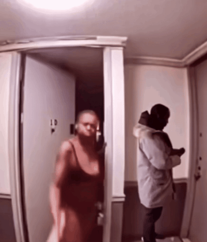 A Woman In A Red Dress Is Walking Into A Room With A Man In A Grey Coat GIF