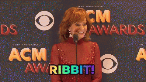 A Woman In A Red Dress Stands In Front Of A Microphone With The Words Acm Awards Behind Her GIF