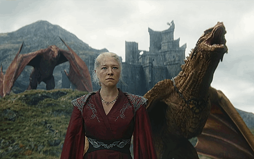 A Woman In A Red Dress Stands Next To A Dragon With A Castle In The Background GIF