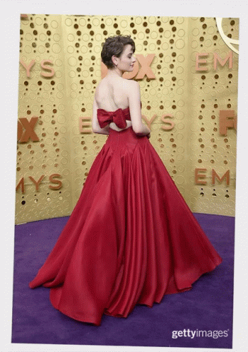 A Woman In A Red Dress Stands On A Purple Carpet In Front Of A Wall That Says Emmys GIF