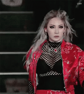 A Woman In A Red Jacket And Black Top Is Standing In Front Of A Staircase . GIF