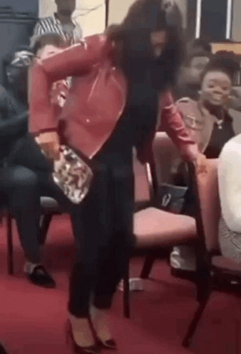 A Woman In A Red Jacket Is Dancing In Front Of A Crowd Of People Sitting In Chairs . GIF