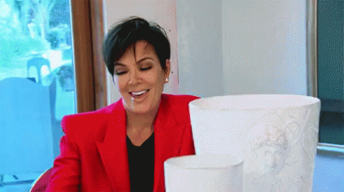 A Woman In A Red Jacket Is Smiling While Sitting At A Table With Two Vases . GIF