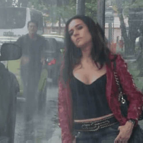 a woman in a red jacket is standing in the rain in front of a bus stop .