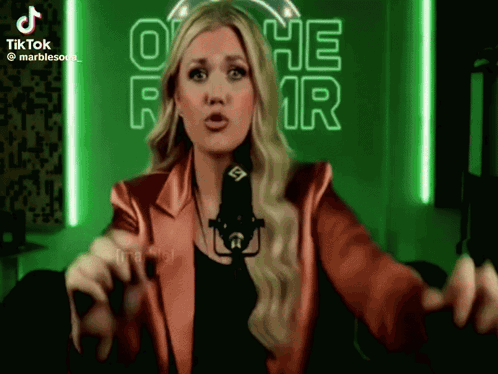 A Woman In A Red Jacket Stands In Front Of A Microphone In Front Of A Neon Sign That Says O He R GIF