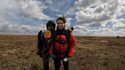 A Woman In A Red Jacket Stands Next To A Man In A Black Jacket Giving A Thumbs Up GIF