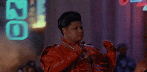A Woman In A Red Leather Jacket Is Standing In Front Of A Neon Sign That Says ' J ' On It GIF