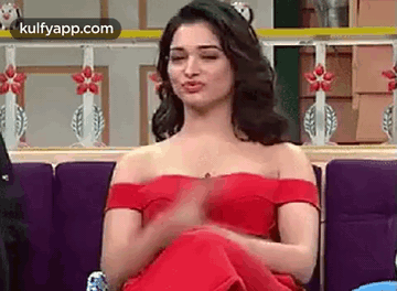 A Woman In A Red Off The Shoulder Dress Is Sitting On A Couch Making A Funny Face . GIF