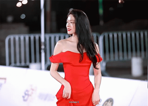 A Woman In A Red Off The Shoulder Dress Stands On A White Carpet GIF