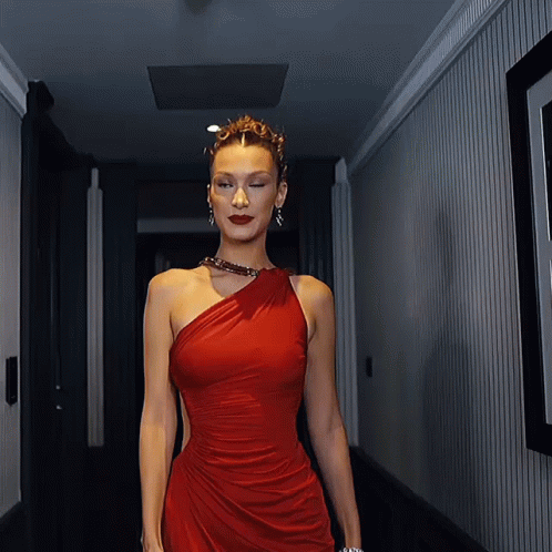 A Woman In A Red One Shoulder Dress Walks Down A Hallway GIF