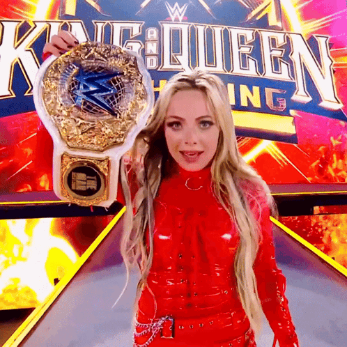A Woman In A Red Outfit Is Holding A Wrestling Championship Belt In Front Of A King Queen Sign GIF