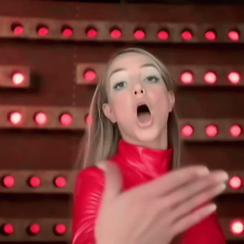 A Woman In A Red Outfit Is Making A Funny Face In Front Of A Wall Of Lights . GIF
