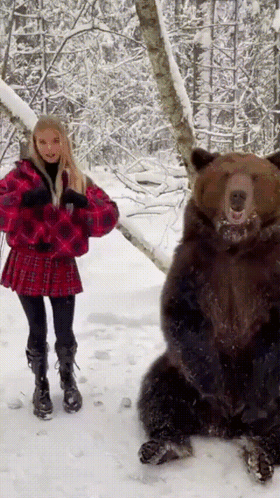 A Woman In A Red Plaid Skirt Stands Next To A Bear In The Snow GIF