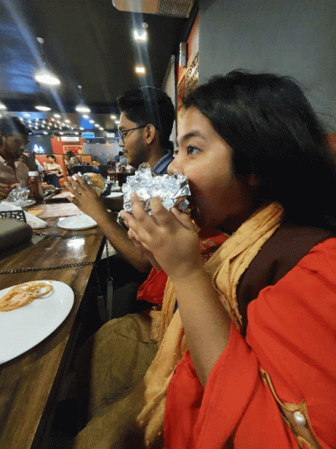 A Woman In A Red Shirt Is Eating A Wrapped Sandwich In A Restaurant GIF