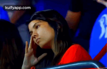 A Woman In A Red Shirt Is Sitting In A Stadium With Her Hand On Her Forehead . GIF