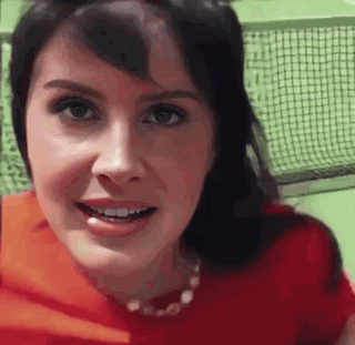 A Woman In A Red Shirt Is Smiling And Looking At The Camera While Standing In Front Of A Tennis Net . GIF