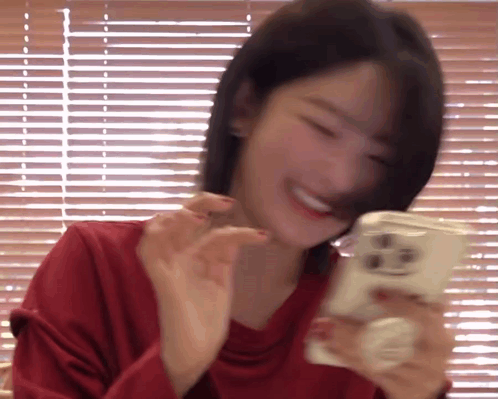 A Woman In A Red Shirt Is Smiling While Holding A Cell Phone In Front Of A Window With Blinds . GIF