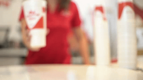 A Woman In A Red Shirt Is Standing Behind A Counter Holding A Cup Of Milkshake . GIF