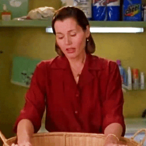 A Woman In A Red Shirt Is Washing Clothes In A Laundry Room With A Bag Of Cheer On The Shelf Behind Her GIF