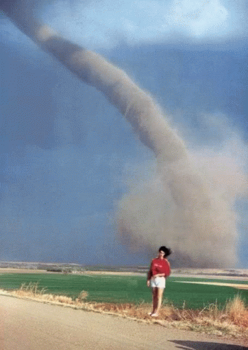 A Woman In A Red Shirt Stands In Front Of A Large Tornado GIF