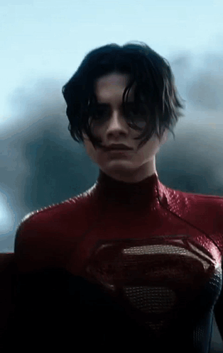 A Woman In A Red Superman Costume Is Standing In Front Of A Window And Looking At The Camera . GIF