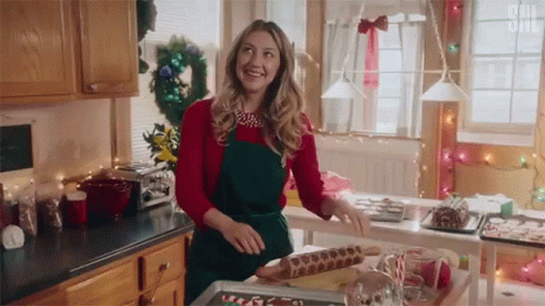 A Woman In A Red Sweater And Green Apron Is Standing In A Kitchen Preparing Christmas Cookies . GIF