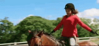 A Woman In A Red Sweater Is Riding A Brown Horse In A Fenced In Area . GIF