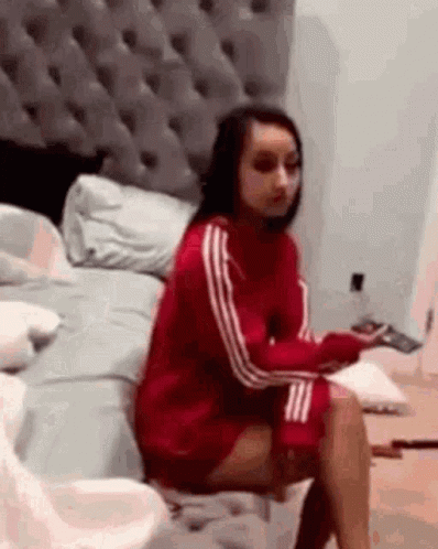 A Woman In A Red Sweater Is Sitting On A Bed With Her Legs Crossed . GIF