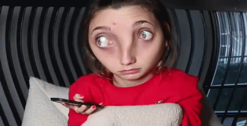 A Woman In A Red Sweater Is Sitting On A Couch Holding A Cell Phone And Making A Funny Face . GIF