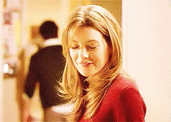 A Woman In A Red Sweater Is Smiling With Her Eyes Closed And A Man In A Black Shirt Is Walking Behind Her . GIF