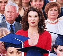 A Woman In A Red Sweater Is Standing In Front Of A Crowd Of People Wearing Graduation Caps . GIF