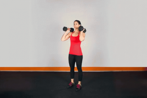 A Woman In A Red Tank Top Is Holding A Pair Of Dumbbells Over Her Head GIF