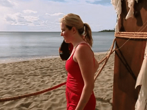 A Woman In A Red Tank Top Stands On A Beach GIF