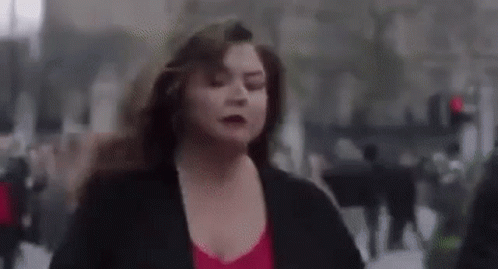 A Woman In A Red Top And Black Jacket Is Running Down A City Street . GIF
